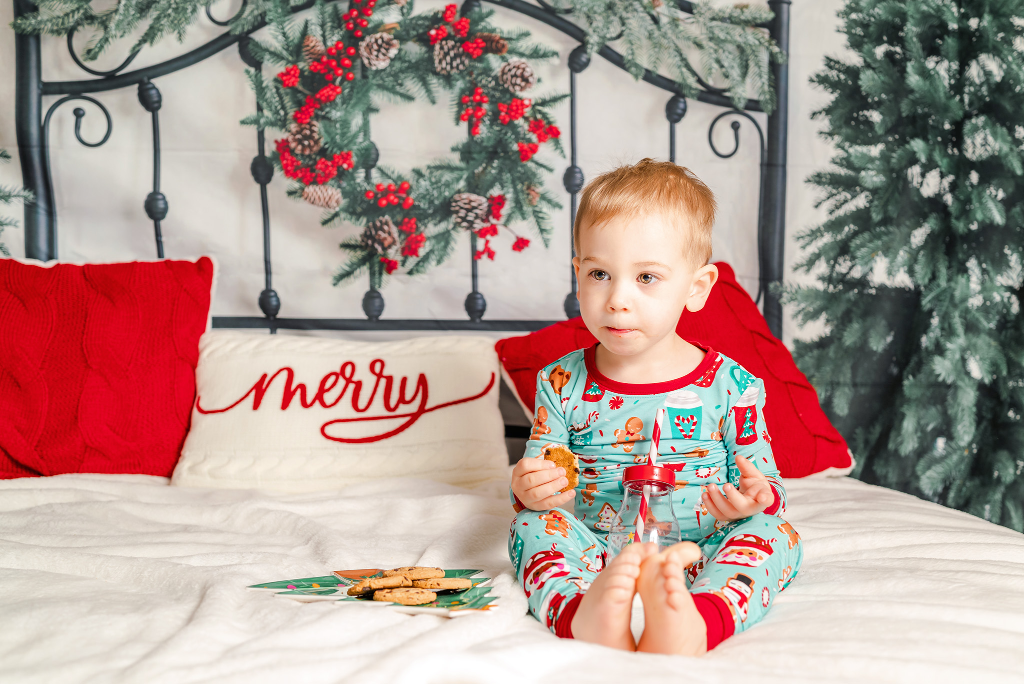 A toddler in Christmas jammies sits on a bed enjoying milk and cookies