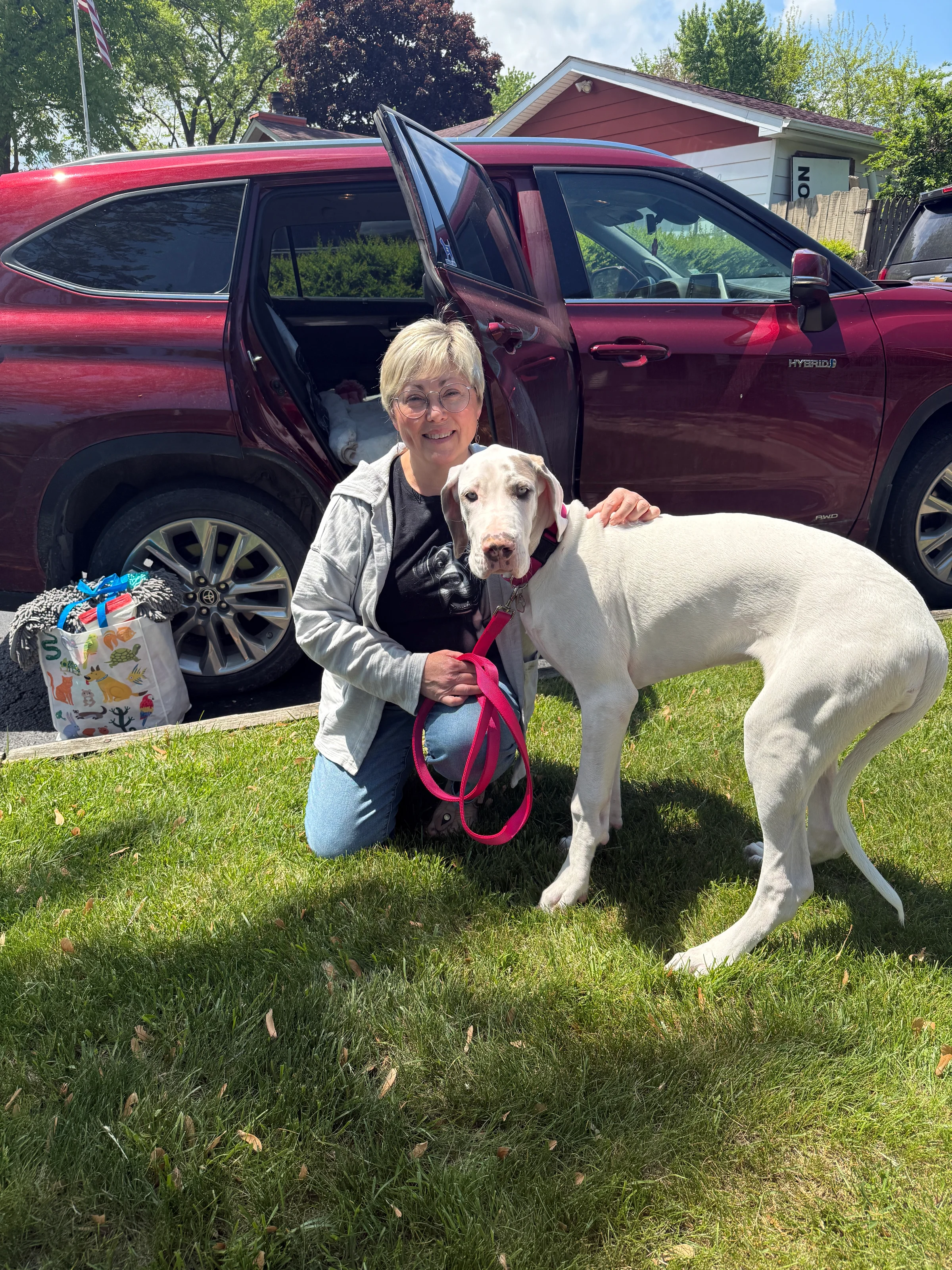 A woman and her dog smile for a photo in front of a bright red car parked on a sunny day.