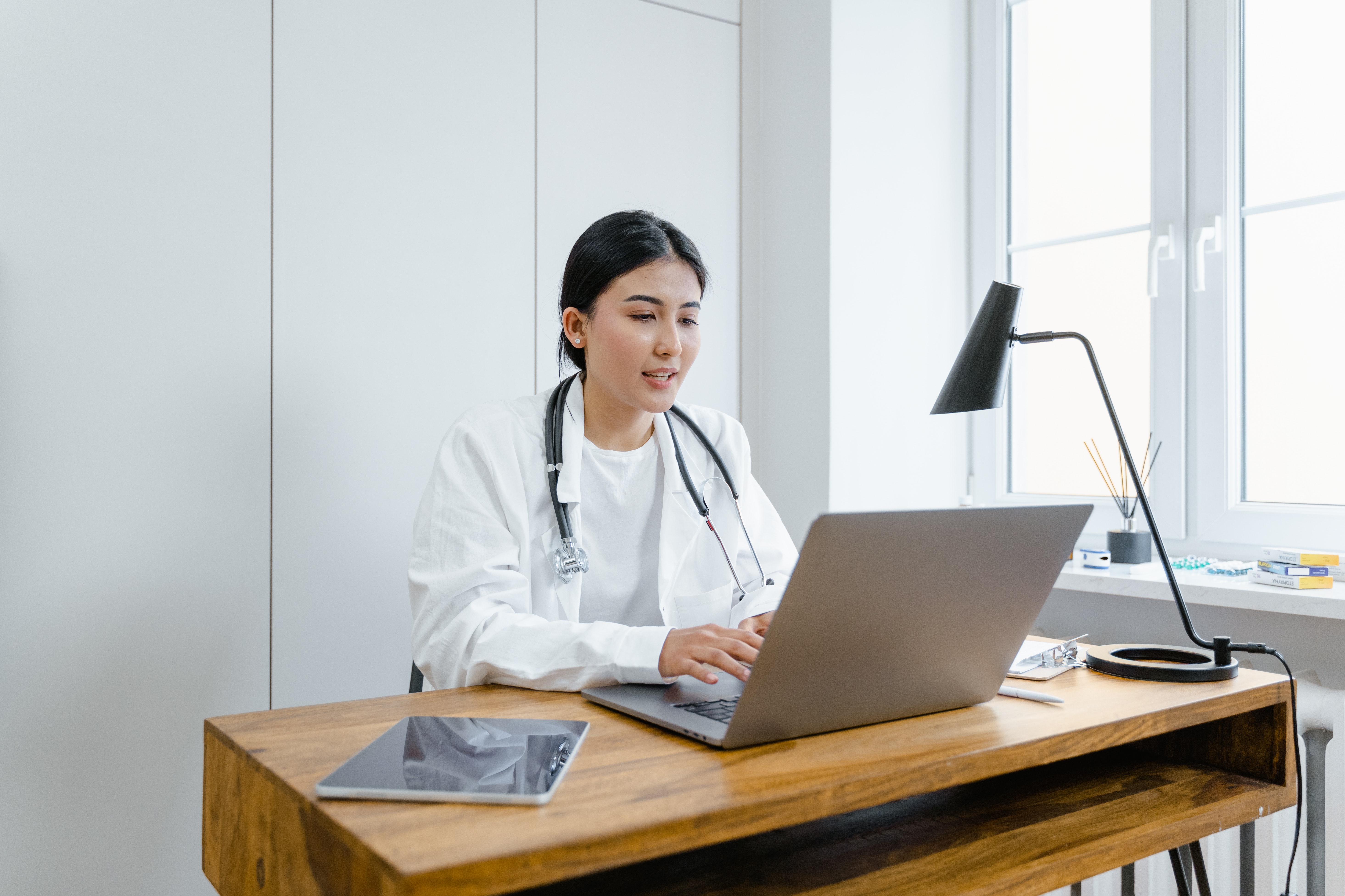 Asian-heritage female doctor in lab coat, sitting at a desk in a minimal office working on a laptop.