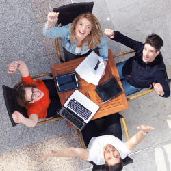 Group of four students with paper and a laptop raising their hands and looking upwards.