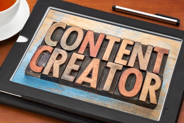 Wooden letter blocks spelling 'CONTENT CREATOR' on a tablet, with a pen and cup of tea in the background, highlighting the concept of digital content creation. Wooden letter blocks spelling 'CONTENT CREATOR' on a tablet, with a pen and cup of tea in the background, highlighting the concept of digital content creation.