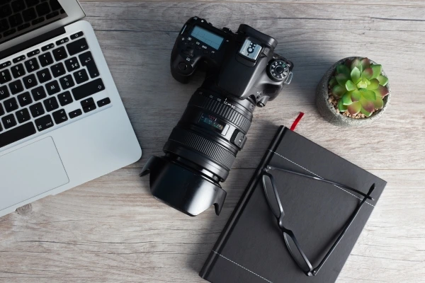 Top view of a photographer's workspace with a laptop, DSLR camera with lens, glasses, notebook, and a succulent plant on a wooden desk, representing creativity and digital content creation. Top view of a photographer's workspace with a laptop, DSLR camera with lens, glasses, notebook, and a succulent plant on a wooden desk, representing creativity and digital content creation.