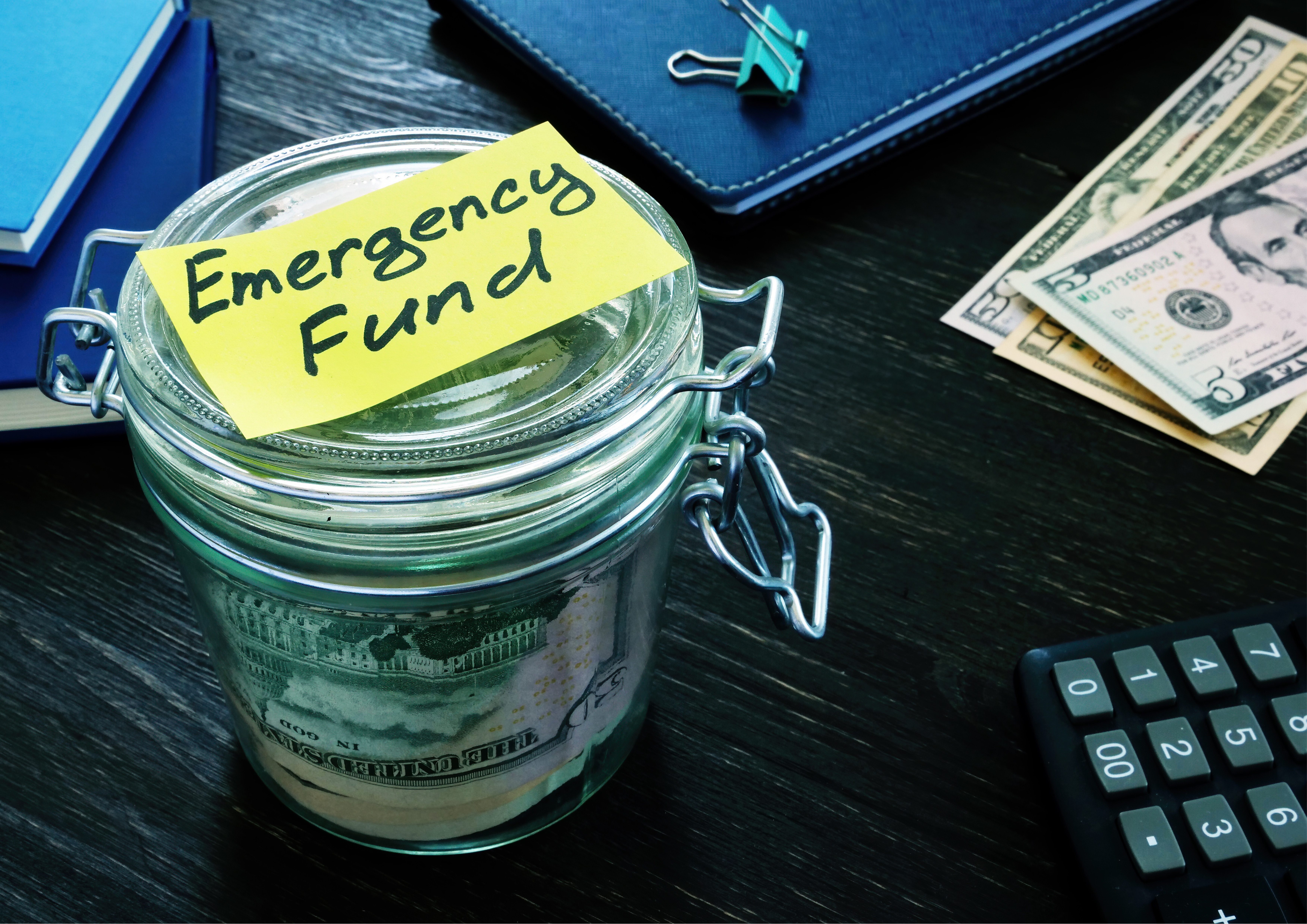 Glass jar labeled "Emergency Fund" filled with U.S. dollar bills, placed on a dark wooden desk with a calculator, cash, and notebooks — symbolizing personal savings and financial planning Glass jar labeled "Emergency Fund" filled with U.S. dollar bills, placed on a dark wooden desk with a calculator, cash, and notebooks — symbolizing personal savings and financial planning
