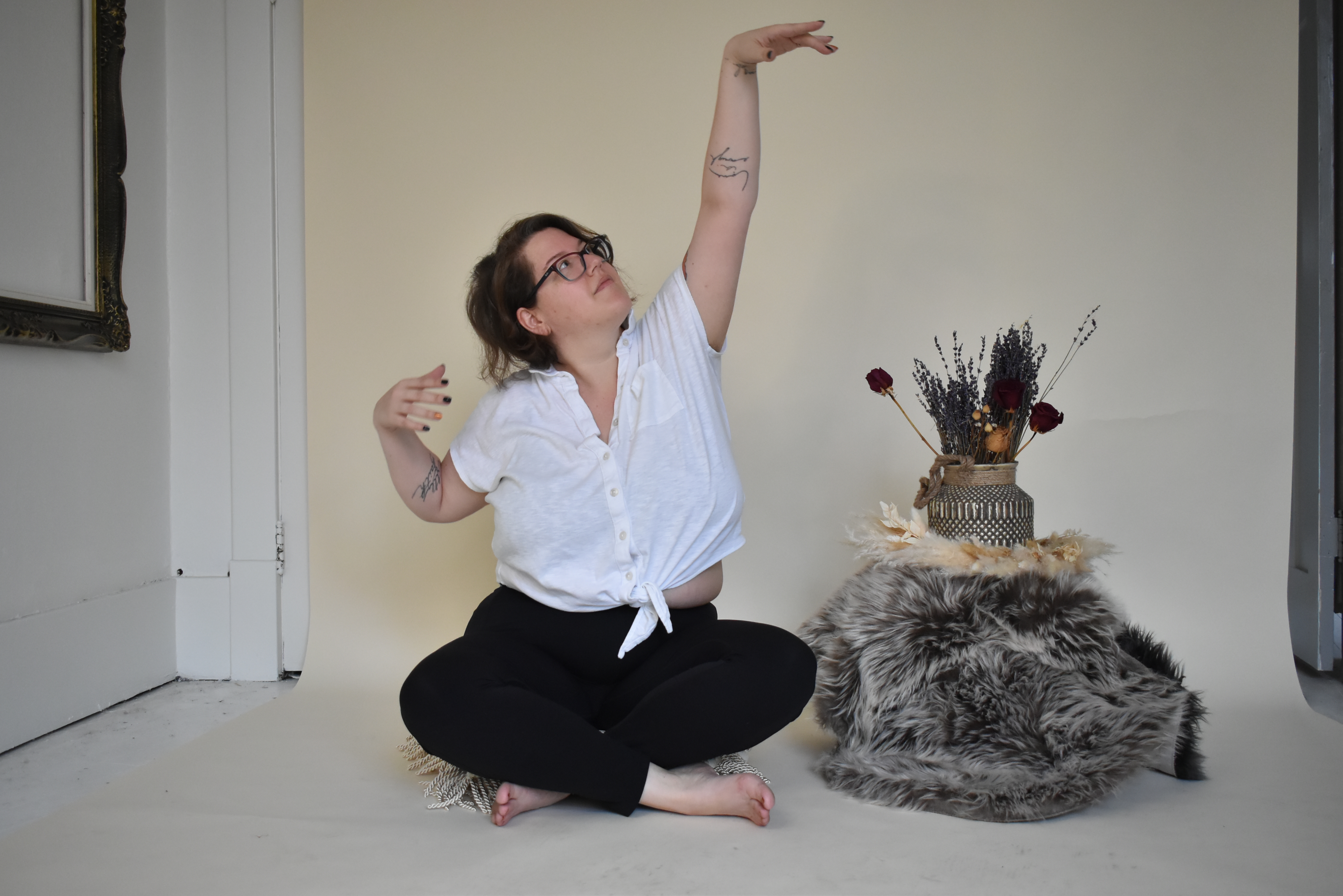 Cheryl sitting crossed legged on the floor in a bright room stretching one hand to the sky and following it with their gaze.