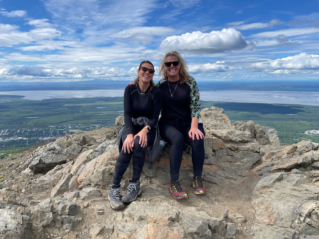 Dr. Barbara and another woman smiling and hugging on a snow-covered mountain peak, dressed in winter hiking gear.