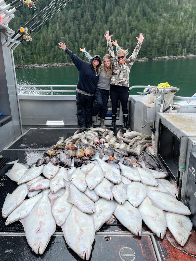 Three women, including Dr. Barbara, standing on a boat deck with arms raised in excitement behind a massive pile of freshly caught fish with a forested shoreline in the background.