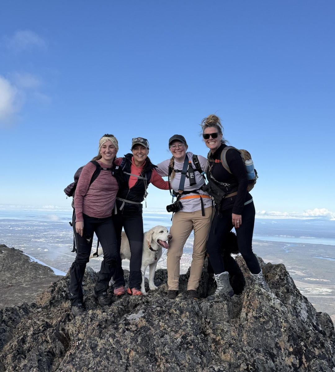 Four women and a dog smiling together on a rocky mountain peak under a clear blue sky, celebrating a successful hike.