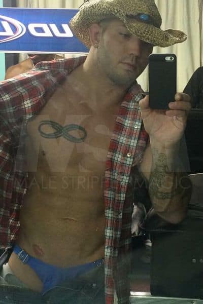 Male stripper Dominic taking a selfie backstage at the male strip club, wearing jeans, a flannel shirt, and a cowboy hat