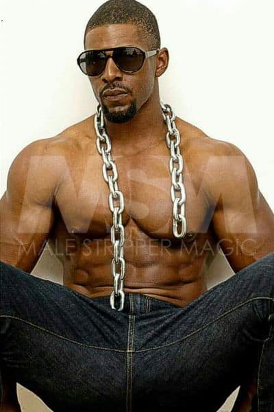 Black male stripper Dream sitting shirtless against the wall, sporting sunglasses, heavy chain and blue jeans