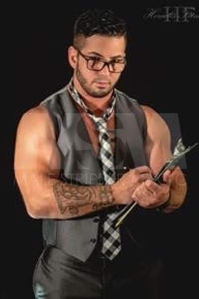 Male stripper Dominic as a captivating teacher, wearing a sleeveless vest, a necktie, and glasses, with a clipboard in hand