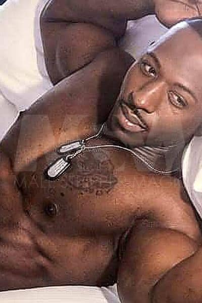 Black male stripper XL lying in bed, dog tags, contemplating the sunny day