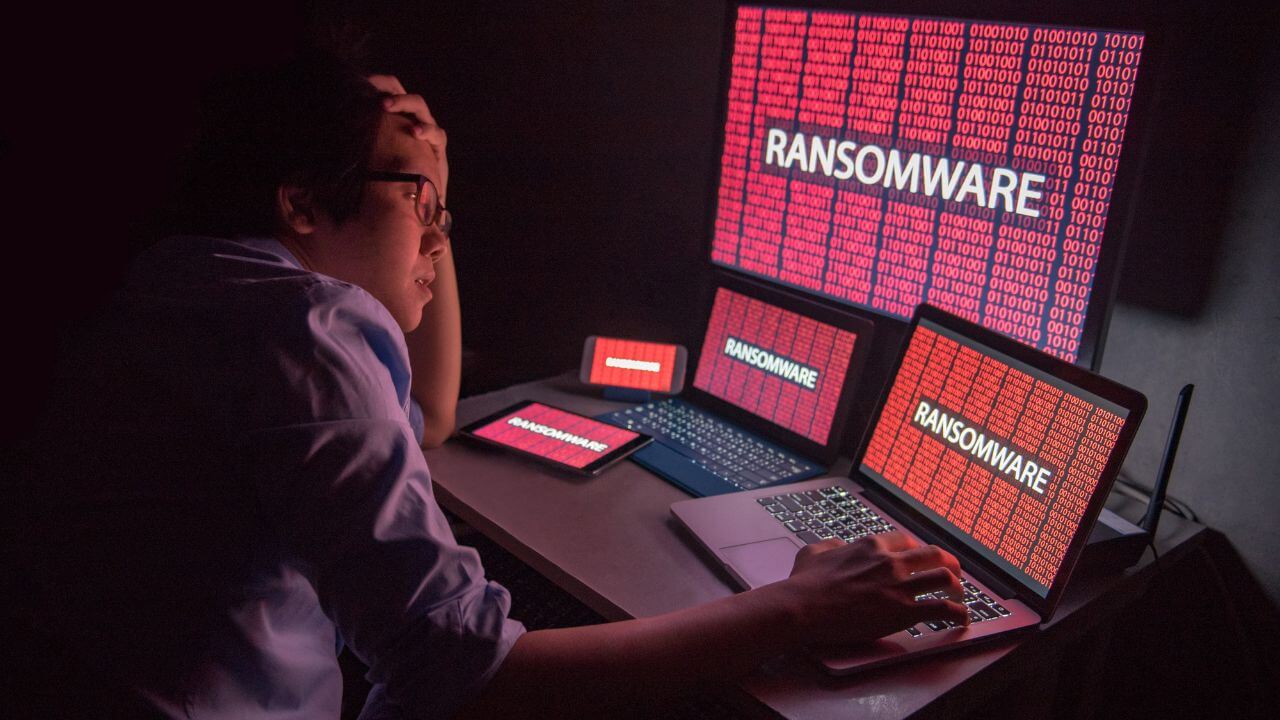 Stressed man surrounded by infected devices displaying ransomware alerts, highlighting the impact of RaaS cyberattacks. Stressed man surrounded by infected devices displaying ransomware alerts, highlighting the impact of RaaS cyberattacks.