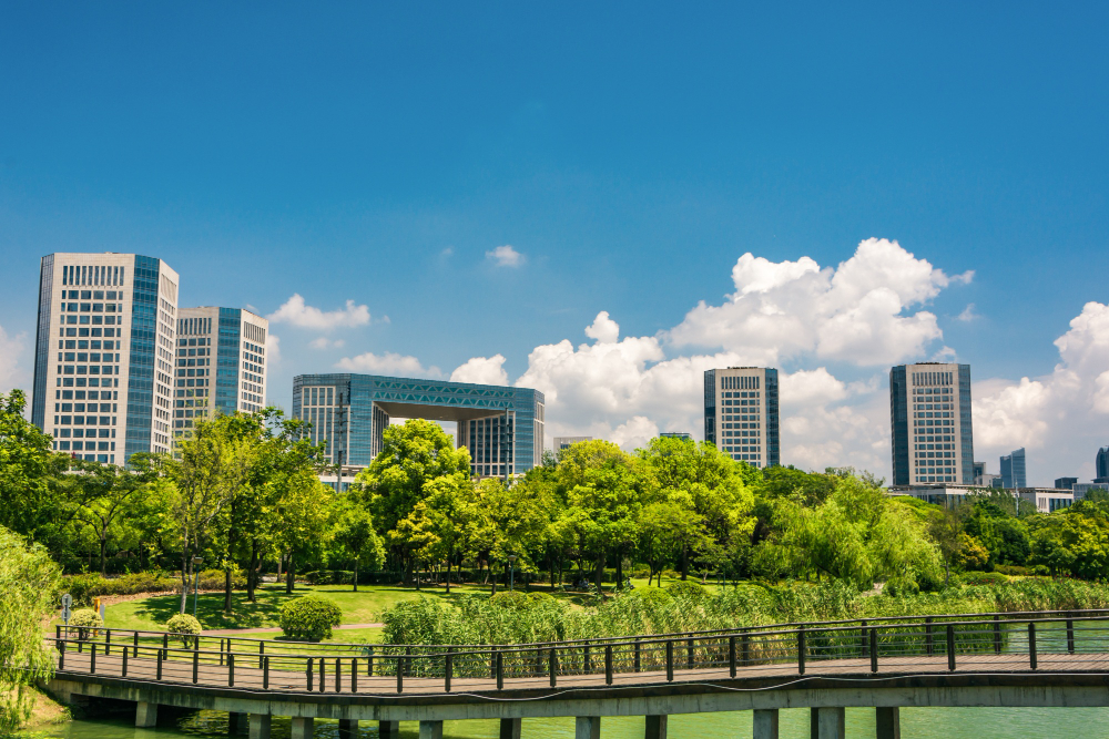 Scenic park view with lush green trees and a wooden bridge over water in the foreground; modern high-rise buildings rise in the background under a clear blue sky with scattered clouds.
