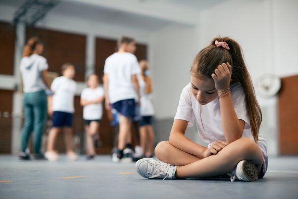 Young girl sitting alone on the gym floor, looking sad and contemplative, while a group of children plays in the background, illustrating feelings of exclusion and loneliness. Young girl sitting alone on the gym floor, looking sad and contemplative, while a group of children plays in the background, illustrating feelings of exclusion and loneliness.