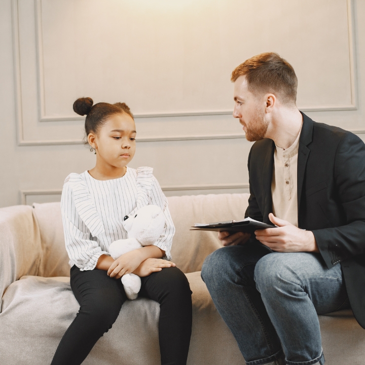 Child sitting with a teddy bear and a concerned expression, talking to an adult male in a blazer, discussing emotional well-being related to financial stress.