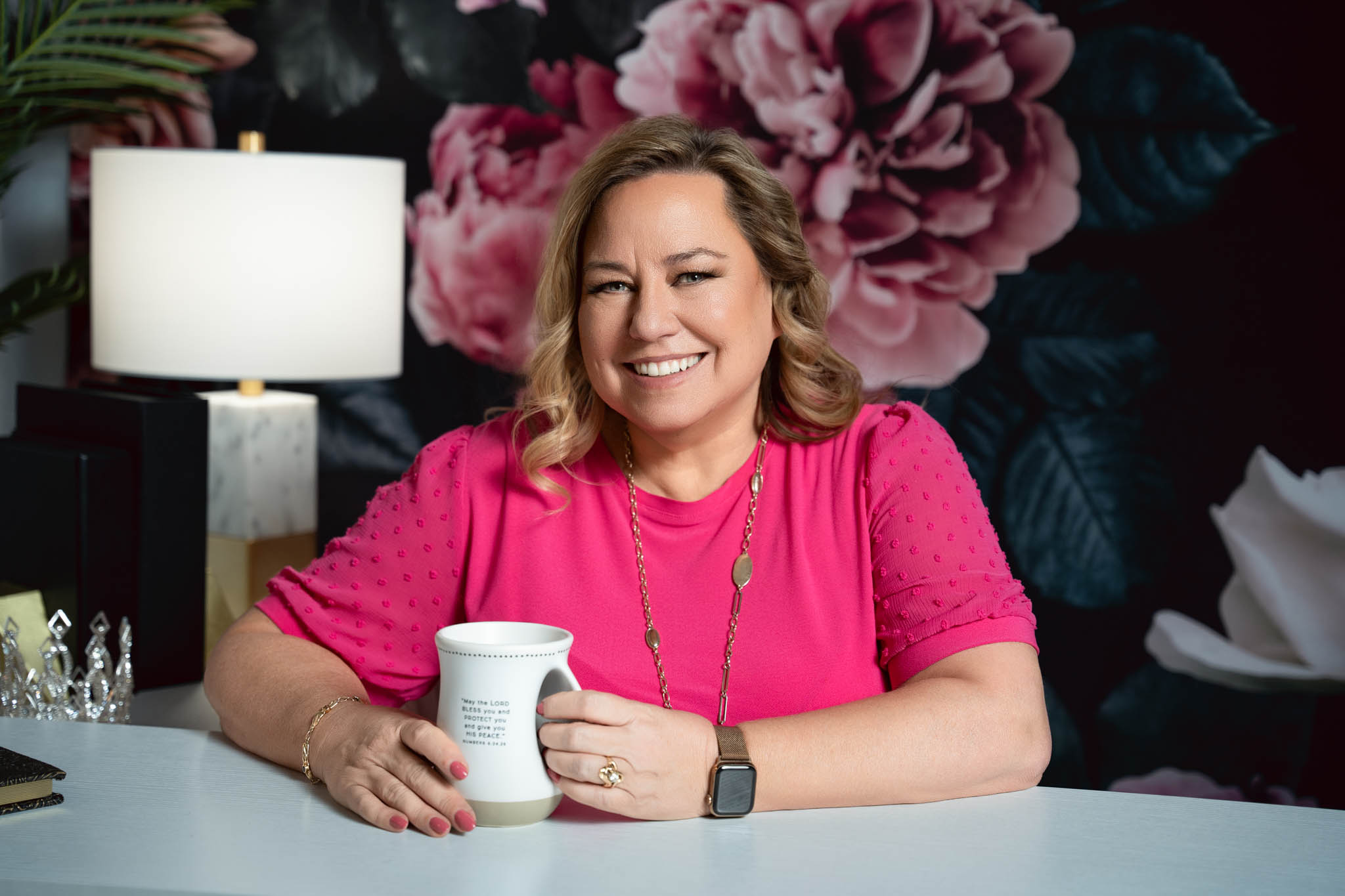 A blonde woman sitting at a white table in a bright pink top, with a floral background holding a coffee cup | Jessica Korff Studios Tucson, AZ