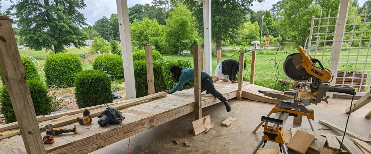 Workers building a wooden accessibility ramp on a back porch surrounded by greenery Workers building a wooden accessibility ramp on a back porch surrounded by greenery