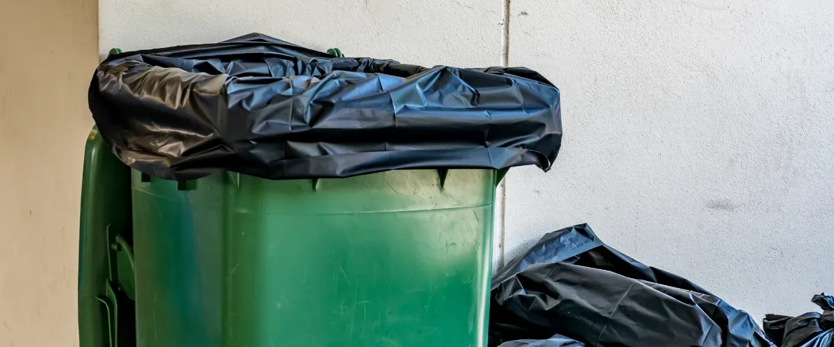 Green trash bin covered with a black garbage bag, with additional full bags on the ground nearby Green trash bin covered with a black garbage bag, with additional full bags on the ground nearby