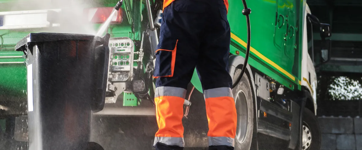 Worker in safety gear pressure washing a black trash bin beside a garbage truck. Worker in safety gear pressure washing a black trash bin beside a garbage truck.