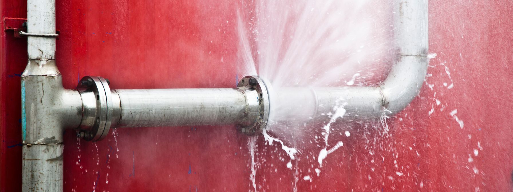 A burst metal pipe sprays water forcefully against a red wall, illustrating a serious plumbing emergency. The image highlights the urgency of addressing pipe failures to prevent flooding and property damage.