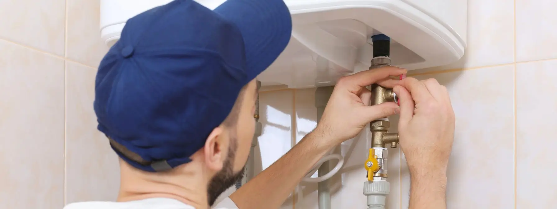 A plumber in a blue cap performs maintenance on a residential water heater, adjusting a valve connection—illustrating professional repair as part of water heater troubleshooting.