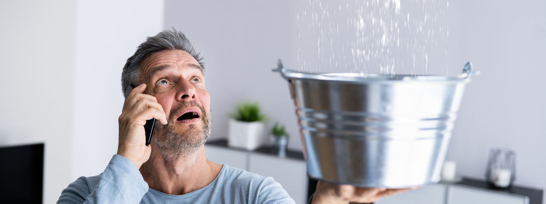 Worried man on the phone holding a metal bucket under a leaking ceiling, trying to catch dripping water during a home plumbing emergency.