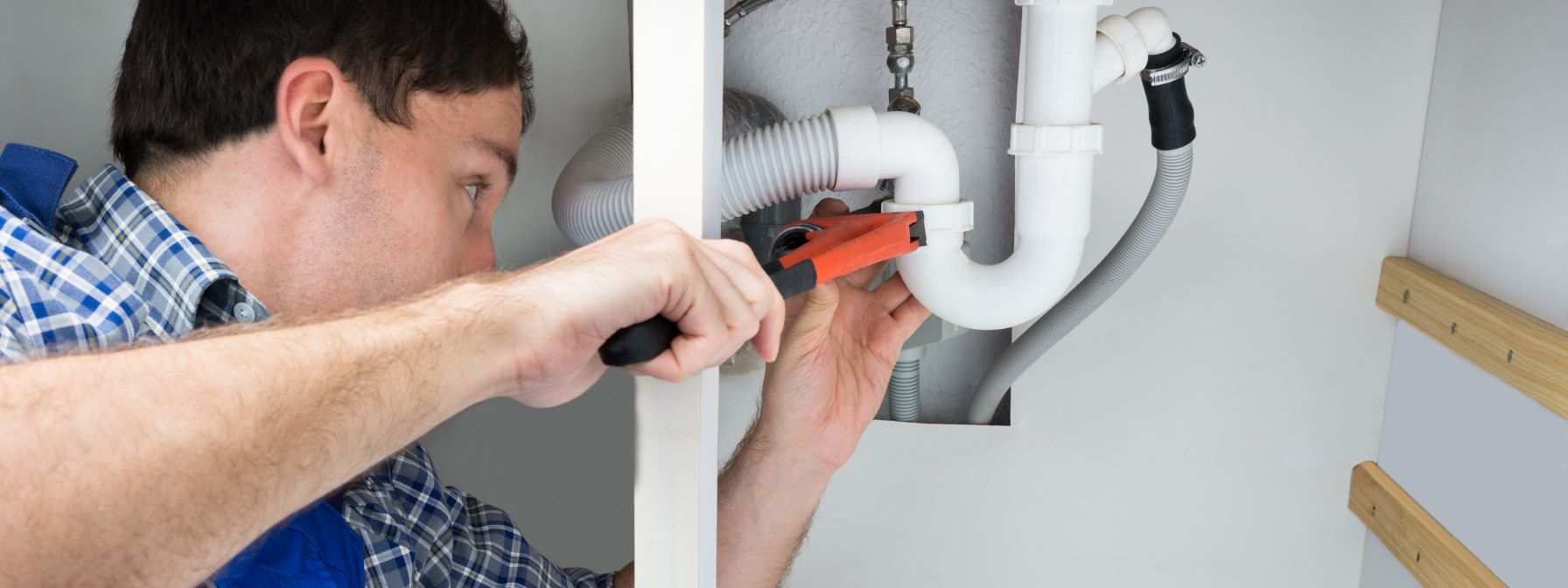 Plumber using a wrench to tighten a white PVC pipe under a sink during an emergency plumbing repair in a residential home.