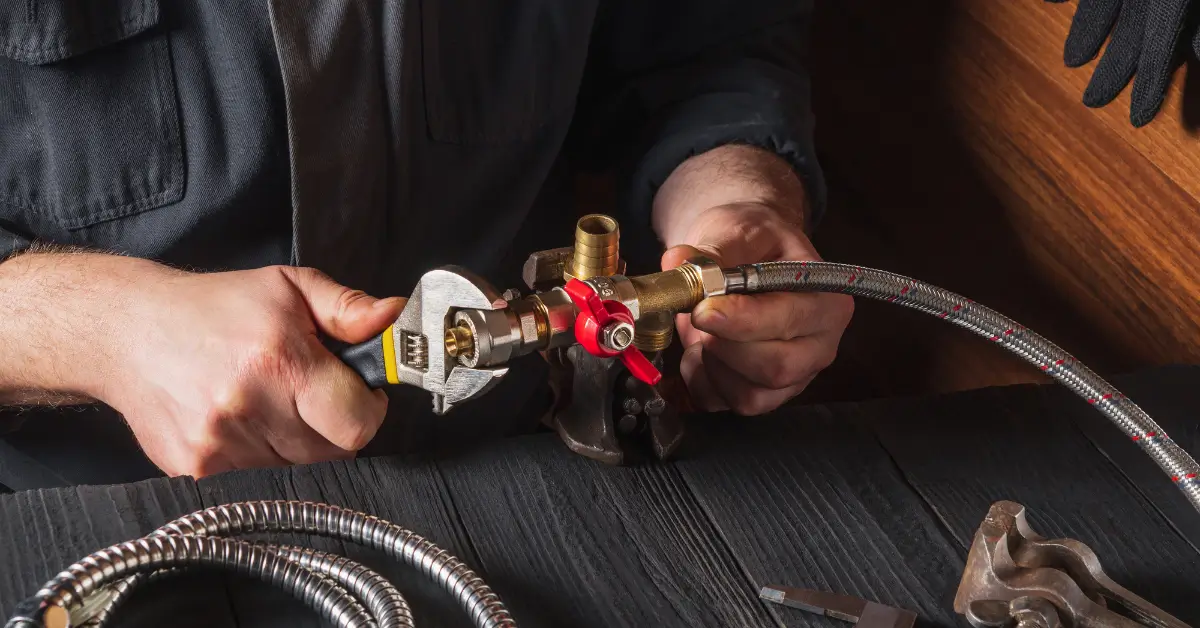 A plumber uses a wrench to tighten a fitting on a flexible braided hose connected to a brass valve with a red handle, while working on a dark wooden surface with tools nearby. A plumber uses a wrench to tighten a fitting on a flexible braided hose connected to a brass valve with a red handle, while working on a dark wooden surface with tools nearby.