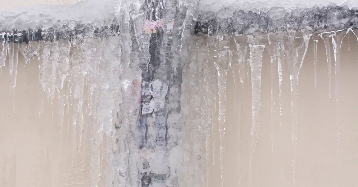 An outdoor pipe is completely covered in thick ice, with long icicles hanging down, indicating it has frozen due to extremely cold temperatures. An outdoor pipe is completely covered in thick ice, with long icicles hanging down, indicating it has frozen due to extremely cold temperatures.