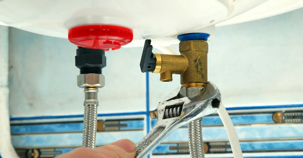 A close-up of a plumber’s hand using a wrench to tighten a connection on a braided metal hose attached to a brass valve under a water heater or tank.