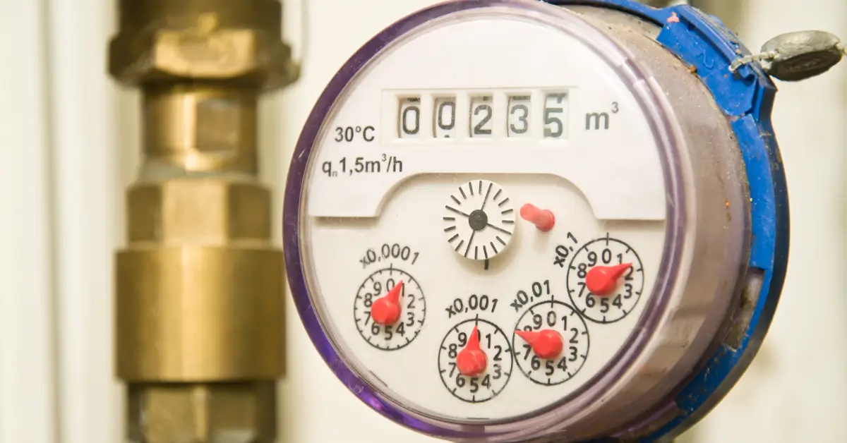 Close-up of a household water meter with a clear plastic cover, showing numeric readings and multiple small dials with red pointers used to measure water consumption.