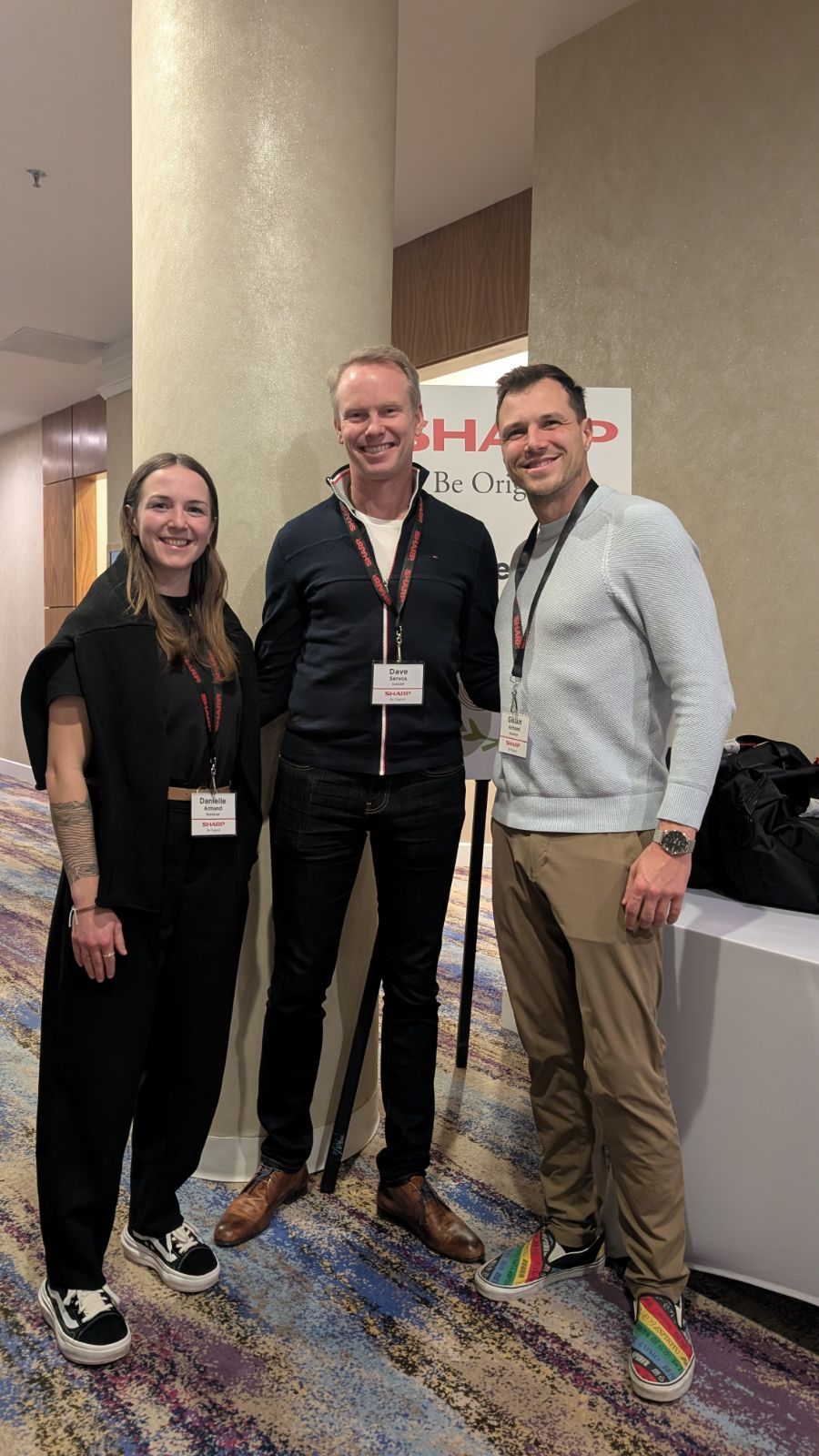 Gislain and Danielle from Randmar stand smiling with a Sharp representative at a business event, wearing name badges and posing in front of a Sharp-branded sign in a conference setting. Gislain and Danielle from Randmar stand smiling with a Sharp representative at a business event, wearing name badges and posing in front of a Sharp-branded sign in a conference setting.