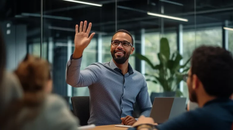 A smiling man in glasses and a blue shirt raises his hand during a business meeting in a modern office, engaging in discussion with colleagues.