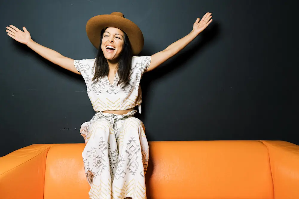 Miki Agrawal sitting on an orange couch and throwing her arms up in wow