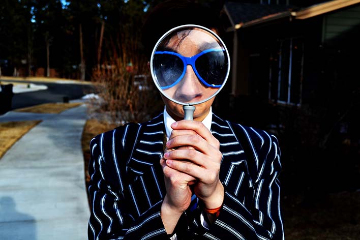 Man's face enlarged while wearing blue sunglasses and blue suit holding magnifying glass in front of face