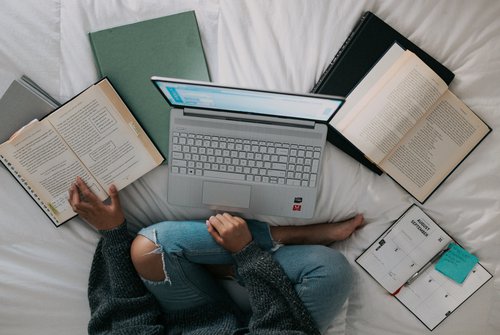 Female student studying, cross-legged on a bed with their laptop and books surrounding them.