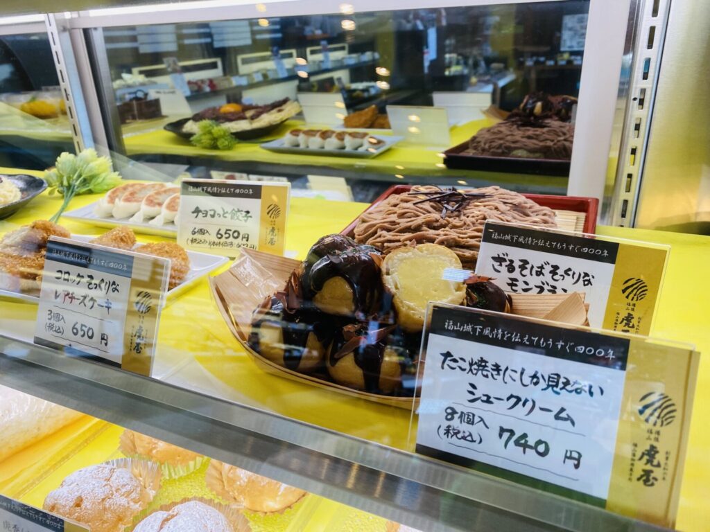 ショーウィンドウの中に並ぶスイーツ「チョコっと餃子」「コロッケそっくりなレアチーズケーキ」「ざるそばそっくりなモンブラン」「たこ焼きにしか見えないシュークリーム」がある