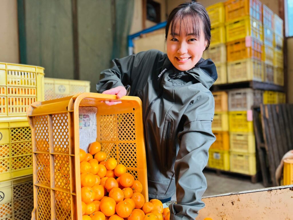 笑顔の善子さんがミカンの入ったケースをもっている様子