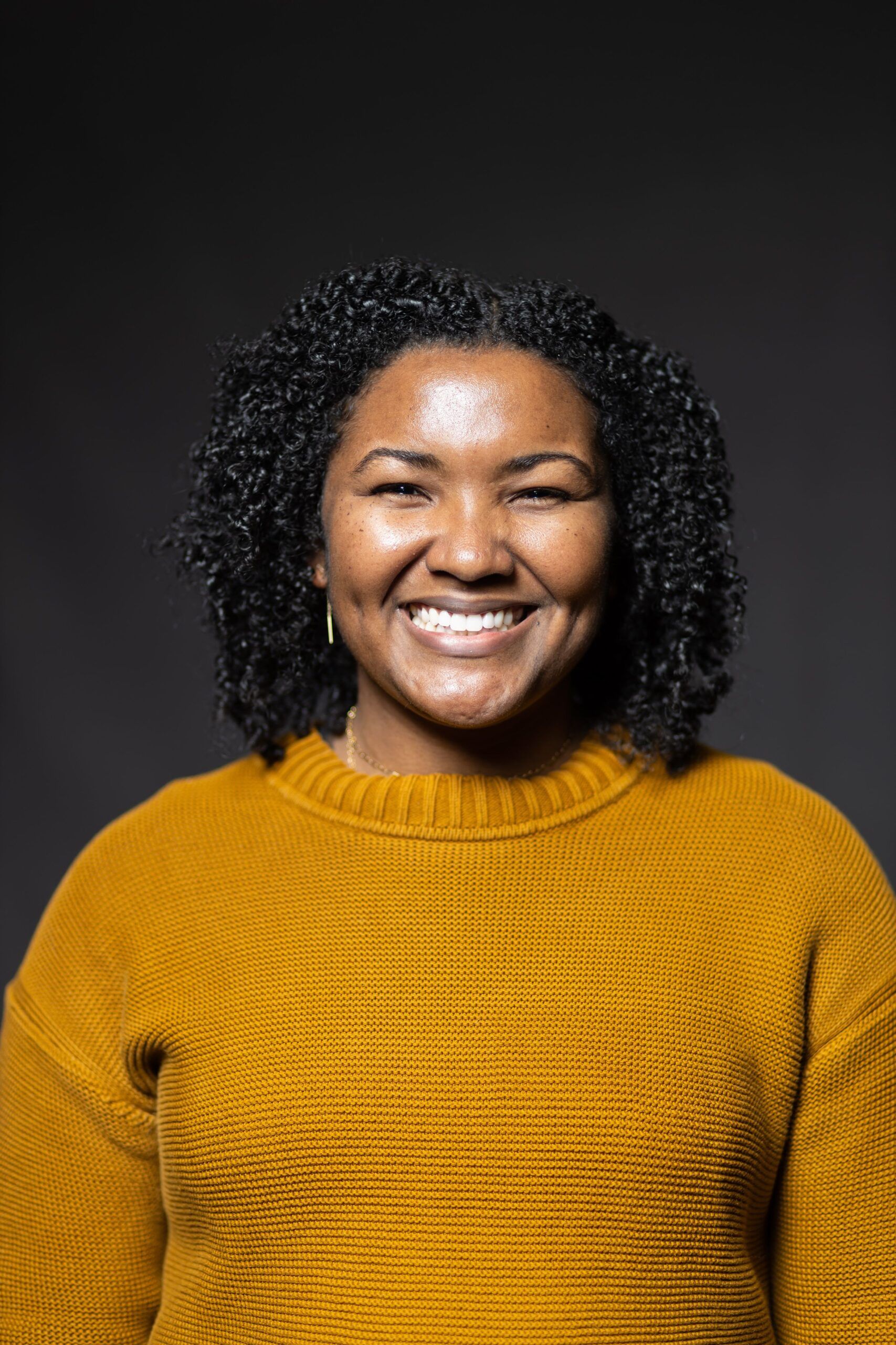 Headshot of Angela with curly, Black hair in a yellow sweater smiling, against a dark backdrop