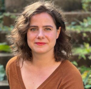 Headshot of Ellen in a brown blouse, with shoulder length brown hair sitting outside