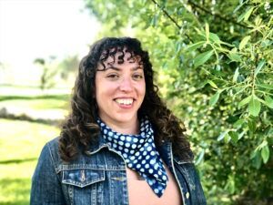 Emily has curly brown, shoulder-length hair, wearing a salmon top with a jean jacket, and blue and white polka dot scarf around her neck