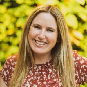 Lindsey has mid-length blone hair, wears a floral shirt, and stands against a natural background.