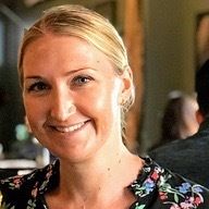 Melissa has blonde hair pulled back into a bun and wears a floral top, smiles at camera.