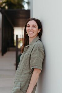 Sarah has long brown hair and is wearing a green jumpsuit, while leaning against a white wall, smiling.