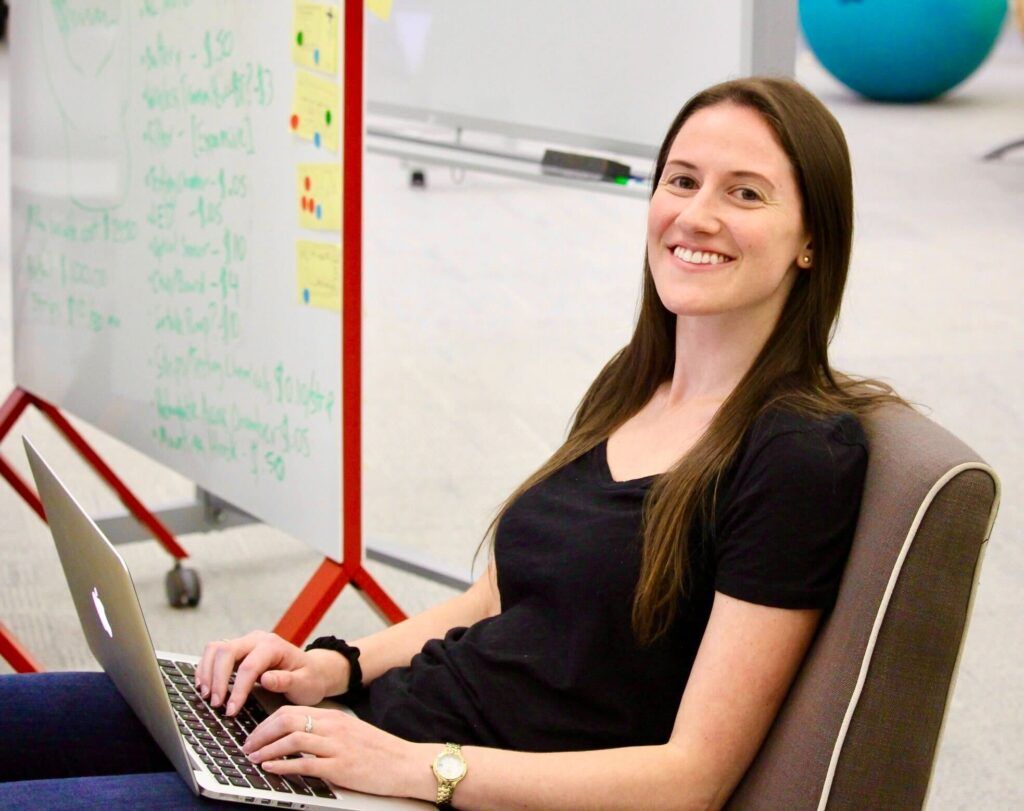 Sam seated in the office with her computer on her lap smiles at the camera