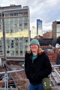 Val wears a teal beanie and black jacket, in front of a city landscape