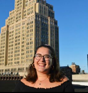 Zoe is in a black shirt, and glasses, with her hair pulled back. A building towers behind her.