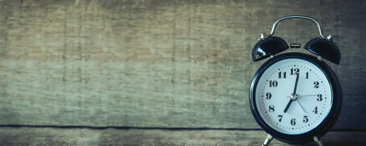 Close-up of a vintage-style black alarm clock with a white clock face, black numerals, and twin bells on top, set against a rustic wooden background.
