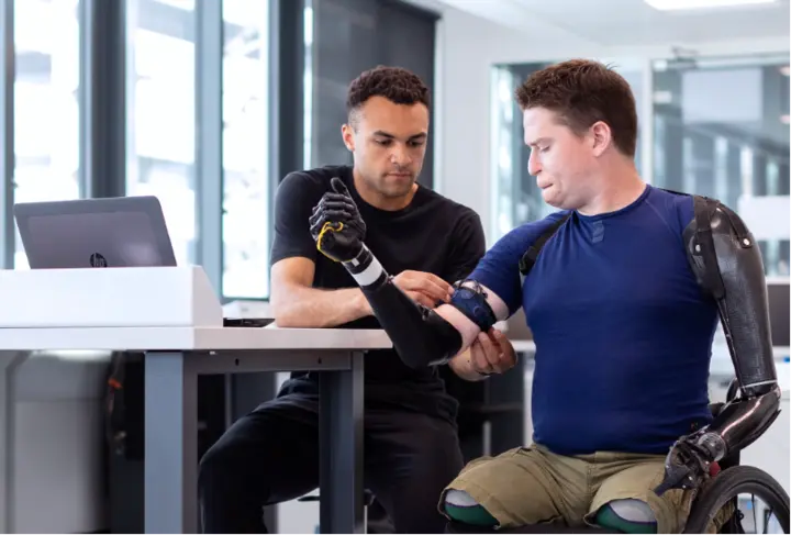 A man adjusting an armband with sensors on the upper arm of another man, who has prosthetic arms and sits in a wheelchair