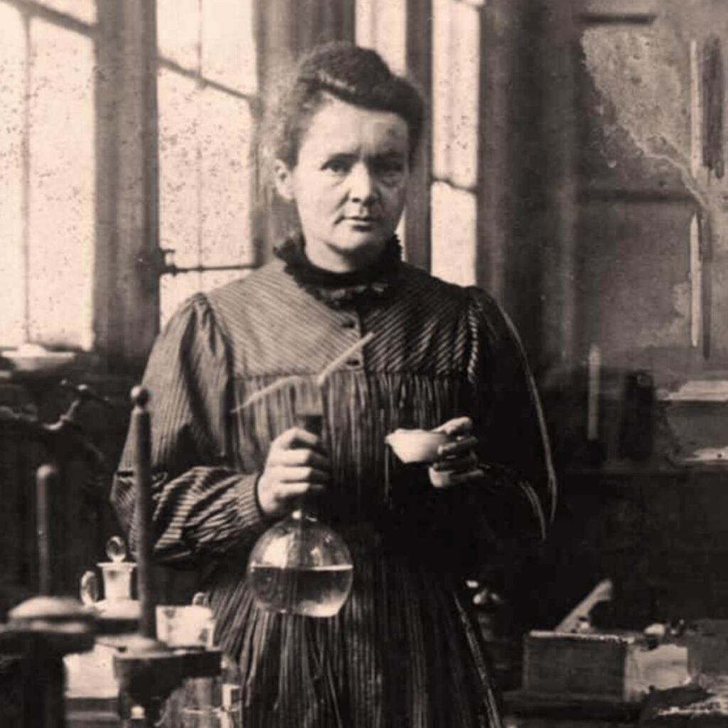 Foto de Marie Curie, a primeira mulher a ganhar um Nobel, em um laboratório segurando um béquer.